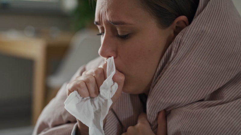 Žena kašlje u maramicu dok sedi umotana u ćebe i deluje iscrpljeno