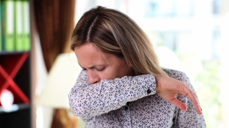 Žena kašlje i pokriva usta rukavom dok stoji u zatvorenom prostoru
