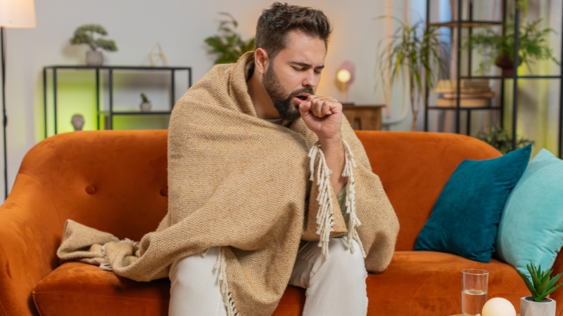 Muškarac umotan u ćebe sedi na kauču i kašlje